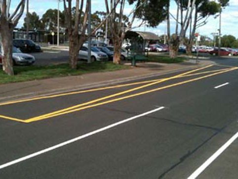 Yellow hatched median markings and a white edge line on a tree-lined road with parked cars beyond
